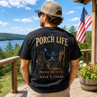 A man stands on a wooden porch with a scenic lake and forest background, wearing a black graphic t-shirt with 'PORCH LIFE' printed on the back. The shirt features a detailed graphic of an outdoor rocking chair scene at night with American flags and the text 'FREEDOM FEELS BETTER ON THE PORCH BACK PORCH APPAREL'.