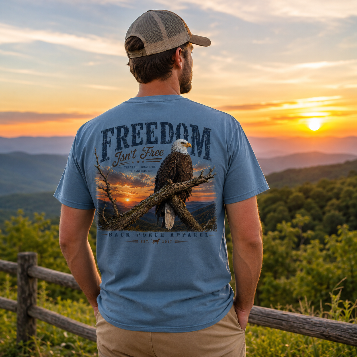 A man stands outdoors at sunset wearing a light blue t-shirt with a large graphic on the back featuring an eagle and the text 'FREEDOM Isn't Free'.