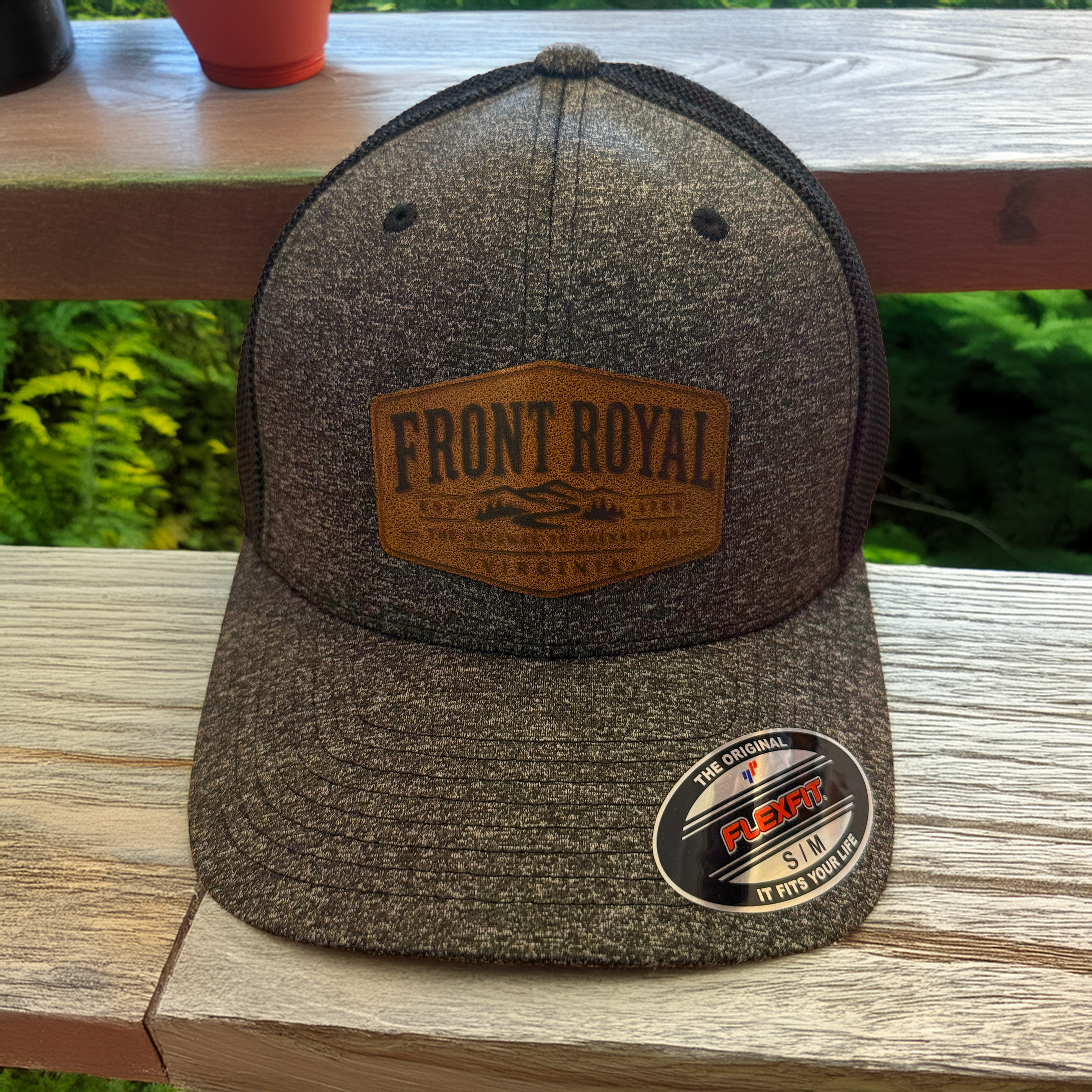 Front Royal Virginia leather patch Flexfit trucker hat displayed on a rustic wooden deck overlooking lush green trees.