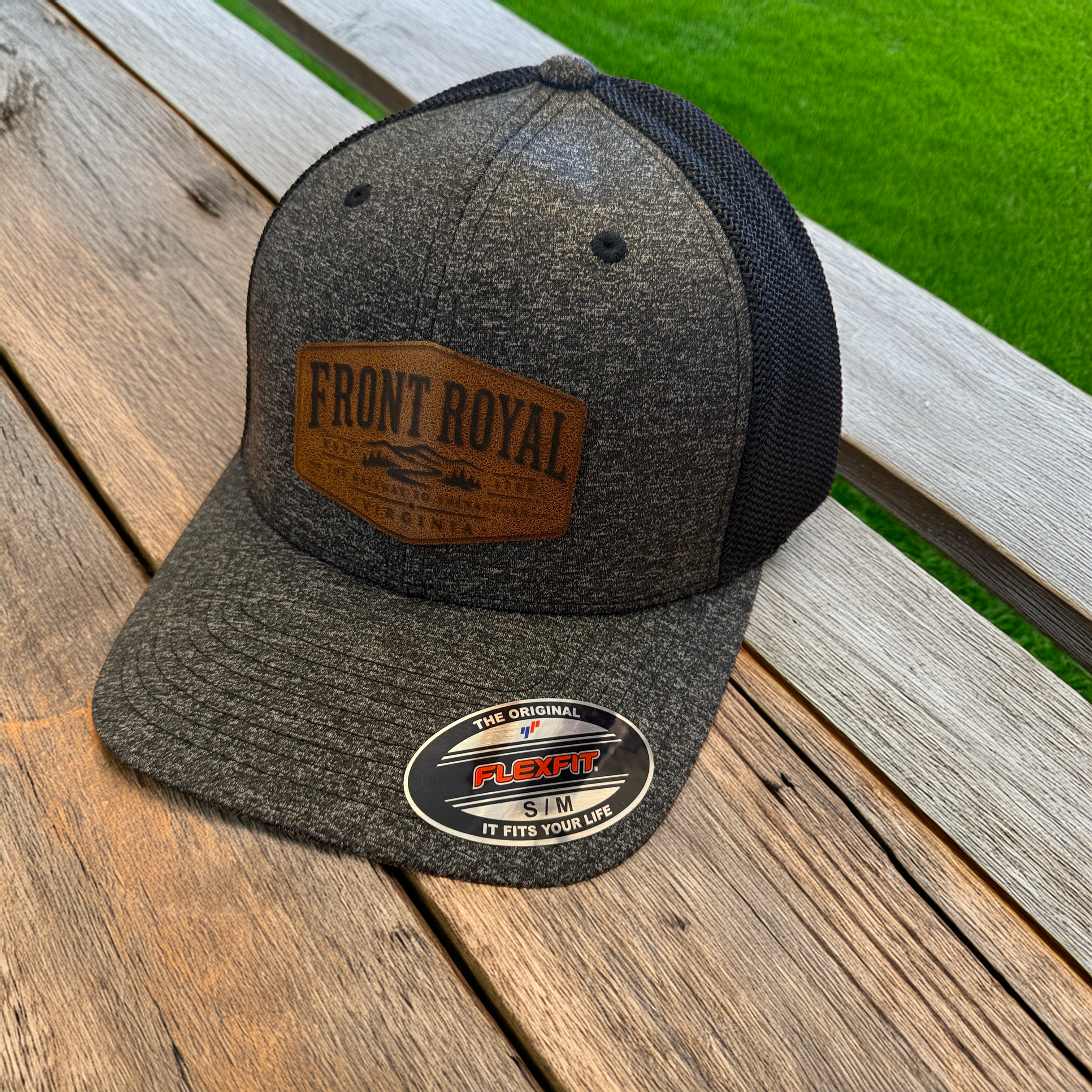 Front Royal Virginia leather patch Flexfit trucker hat resting on a weathered wooden deck with a green lawn in the background.