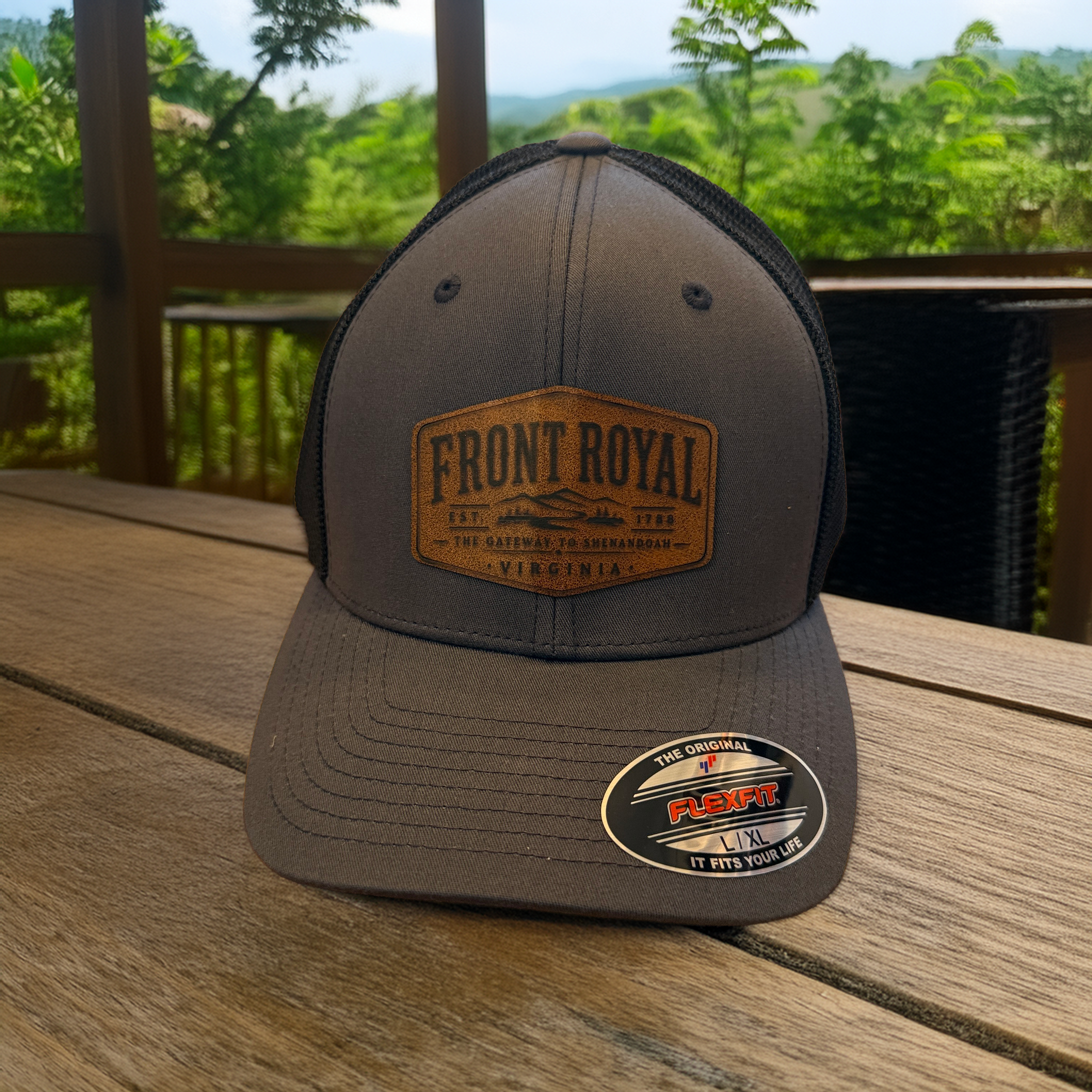 Front Royal Virginia charcoal Flexfit trucker hat with engraved leather patch photographed on a rustic deck overlooking the Shenandoah Valley.