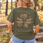 Woman wearing an olive green Simply Southern dragonfly short sleeve T-shirt with wildflowers and God Turns Broken Pieces Into Masterpieces back graphic.