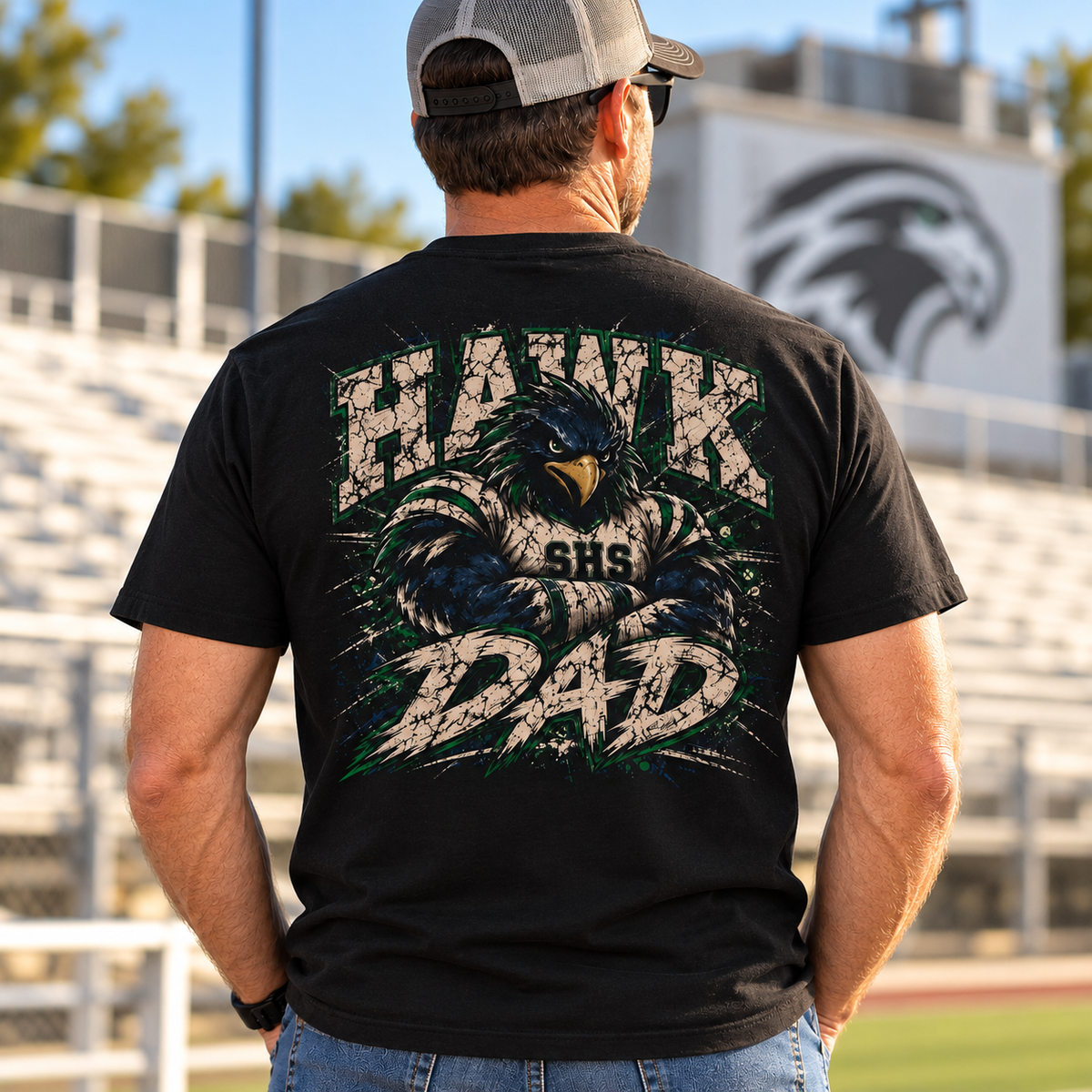 A man viewed from behind wearing a black graphic t-shirt with a large distressed hawk design and the words 'HAWK DAD' on the back, standing on a school field with bleachers and a stadium sign in the background.