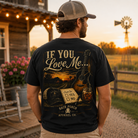 A man stands with his back to the camera wearing a black graphic t-shirt with a rustic country design that reads 'IF YOU Love Me.
