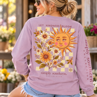 Woman modeling a Wisteria Simply Southern Let Kindness Bloom long sleeve shirt featuring a retro smiling sun and floral back graphic.