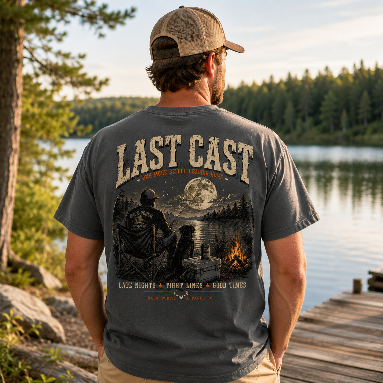 A man standing on a wooden dock by a lake, viewed from behind, wearing a dark gray t-shirt with a large graphic print on the back. The graphic features the text 'LAST CAST' and an illustration of a campfire, moon, and two people sitting in chairs under a starry sky.