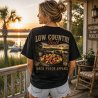 A woman stands on a wooden porch at sunset wearing a black graphic T-shirt with a large back print reading 'LOW COUNTRY Boil' and 'BACK PORCH APPAREL,' showing a pot of shrimp and grits. The background includes water, palm trees, a rocking chair, and a porch sign.
