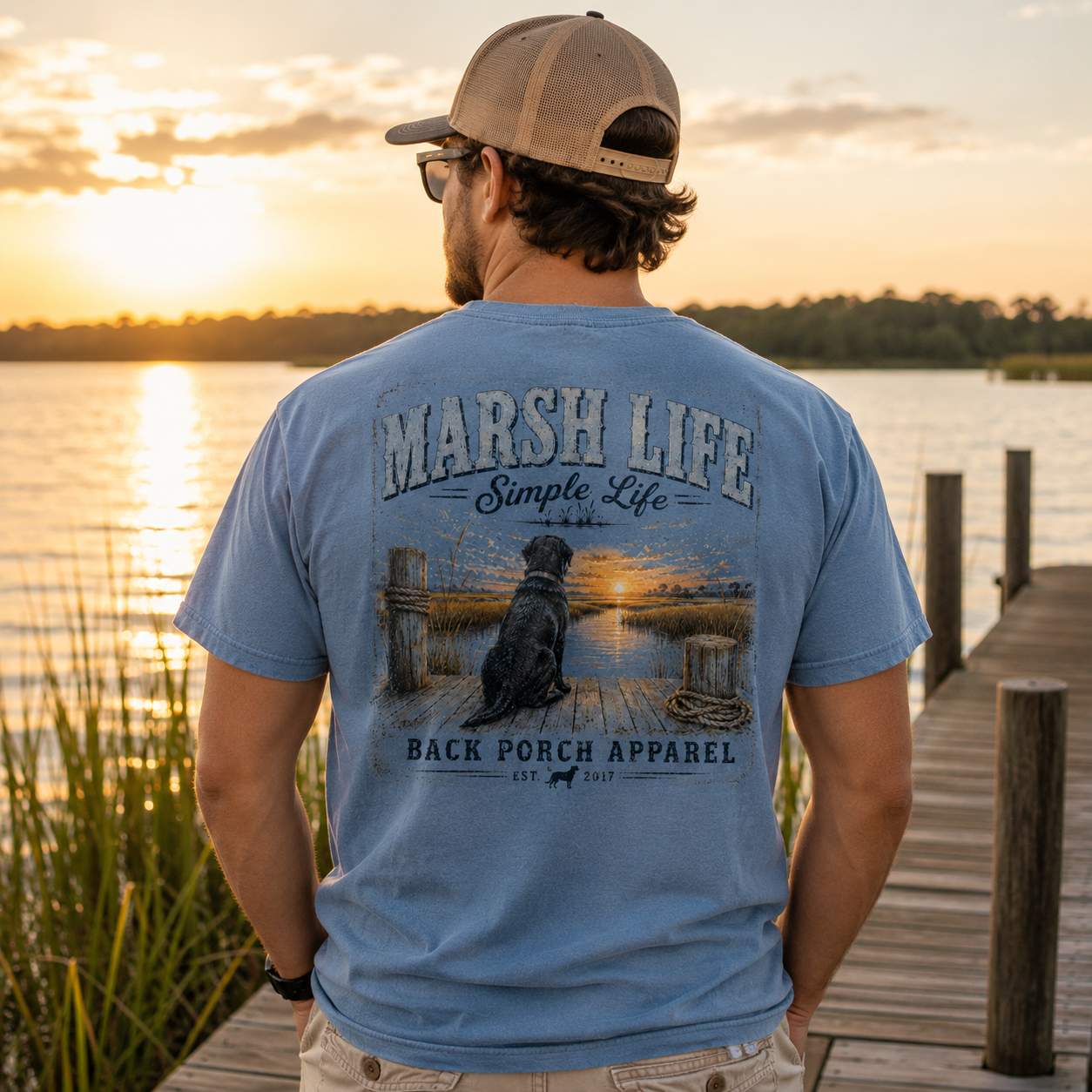 A man standing on a wooden dock at sunset wearing a light blue t-shirt with a large graphic on the back that reads 'MARSH LIFE Simple Life BACK PORCH APPAREL'.