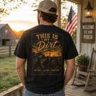 A man stands on a rustic porch wearing a black graphic T-shirt with a large vintage-style tractor and text print reading “THIS IS MY Dirt.” The photo is taken from behind, showing the back of the shirt in a real outdoor rural setting with an American flag, porch light, and wooden cabin in the background.