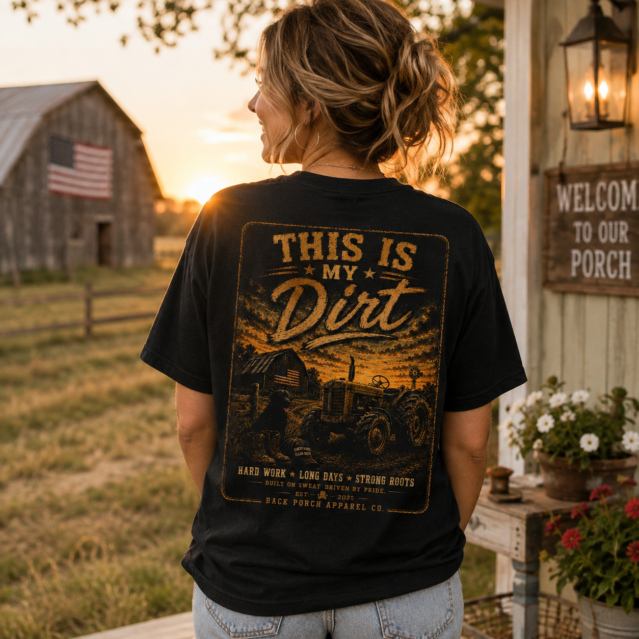 A woman stands on a porch at sunset wearing a black graphic T-shirt with a large distressed yellow-brown print reading 'THIS IS MY Dirt' and tractor artwork. The background shows a barn, American flag, lantern, flowers, and a 'Welcome to Our Porch' sign.