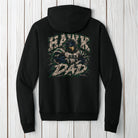 A black pullover hoodie shown from the back, featuring a large graphic print of a hawk with the text 'HAWK DAD' and 'CHG' on the back, displayed against a light gray wood plank background.