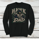 A black crewneck sweatshirt featuring a distressed graphic of a hawk mascot with the text 'HAWK DAD' and 'SHS' on the front, displayed flat against a white wood plank background.