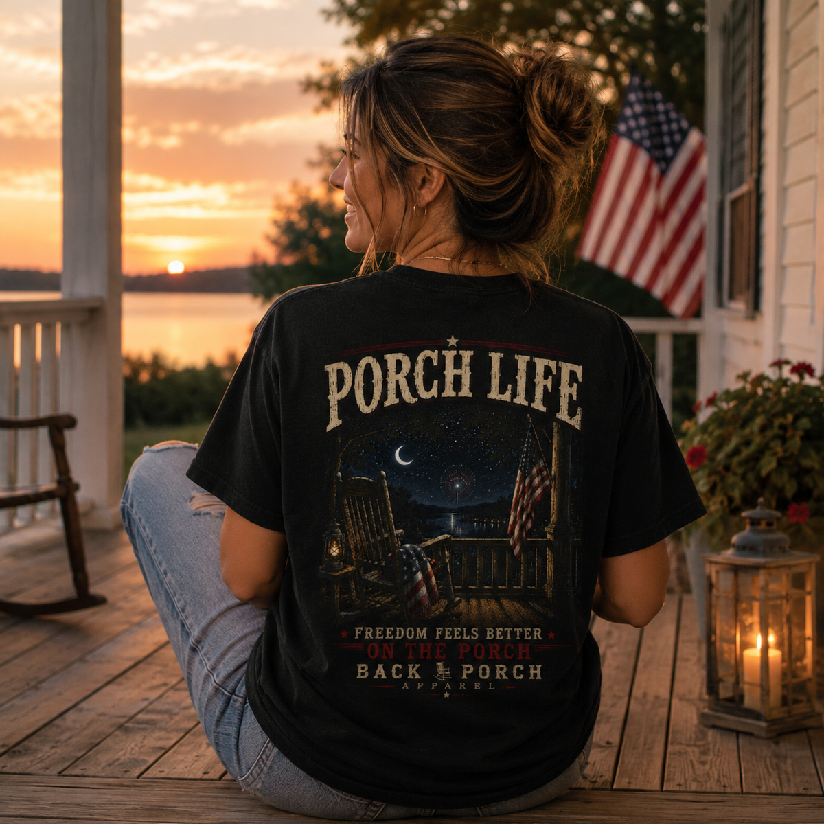 A woman sits on a wooden porch at sunset wearing a black graphic T-shirt with the words “PORCH LIFE” and a patriotic backyard scene printed on the back, paired with light blue jeans. An American flag, lantern, and house are visible in the background.