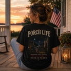 A woman sits on a wooden porch at sunset wearing a black graphic T-shirt with the words “PORCH LIFE” and a patriotic backyard scene printed on the back, paired with light blue jeans. An American flag, lantern, and house are visible in the background.