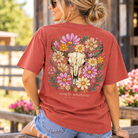 Woman wearing a rust Simply Southern short sleeve T-shirt featuring a Western longhorn skull surrounded by colorful pink, yellow, and cream wildflowers.