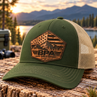 Richardson 112FP loden green and tan mesh trucker hat with light tan BPA American flag mountain leather patch, displayed on rustic wood beside a mountain lake.
