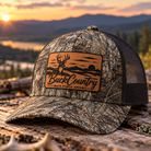A camouflage trucker cap with a brown leather patch reading BuckCountry Back Porch Apparel, resting on a log with a mountain lake sunset background.