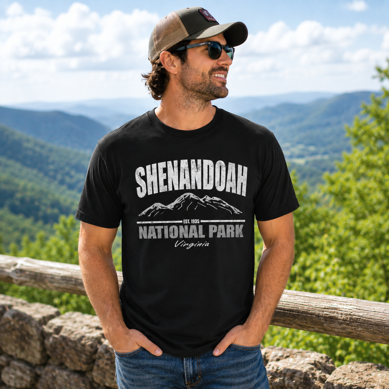 A man wearing a black graphic t-shirt that reads 'SHENANDOAH NATIONAL PARK Virginia' with a mountain illustration, standing outdoors in front of a scenic mountain landscape.