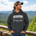 A man wearing a dark gray hoodie with a 'Shenandoah National Park' graphic, standing outdoors in front of a scenic mountain landscape.