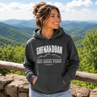 A woman wearing a dark charcoal gray hoodie with a white graphic print that reads 'SHENANDOAH NATIONAL PARK Virginia' stands outdoors in front of a wooden railing, with a scenic mountain landscape in the background.