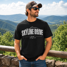 A man wearing a black graphic t-shirt with 'SKYLINE DRIVE Virginia' printed in white, standing outdoors in front of a scenic mountain landscape.