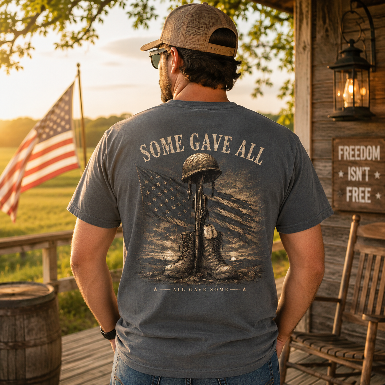 A man viewed from behind wearing a gray graphic t-shirt with a military memorial design on the back, standing on a rustic wooden porch with an American flag in the background.