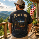 A man stands on a wooden porch wearing a dark graphic T-shirt with the words “PORCH LIFE” and a porch scene printed on the back. The shirt also reads “THE STARS ARE BETTER ON THE PORCH BACK PORCH APPAREL.