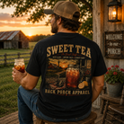 A man wearing a black graphic t-shirt with a large 'Sweet Tea' design on the back, sitting on a porch at sunset holding an iced tea mug.