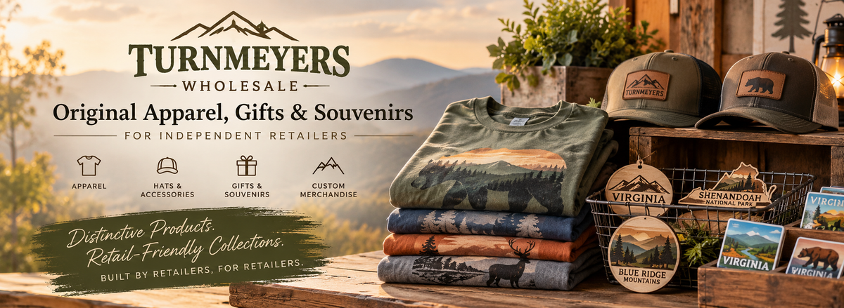 Turnmeyers Wholesale apparel, gifts and souvenirs for independent retailers, featuring outdoor graphic T-shirts, leather-patch hats, Virginia magnets, and Shenandoah National Park ornaments against a scenic Blue Ridge Mountains backdrop.