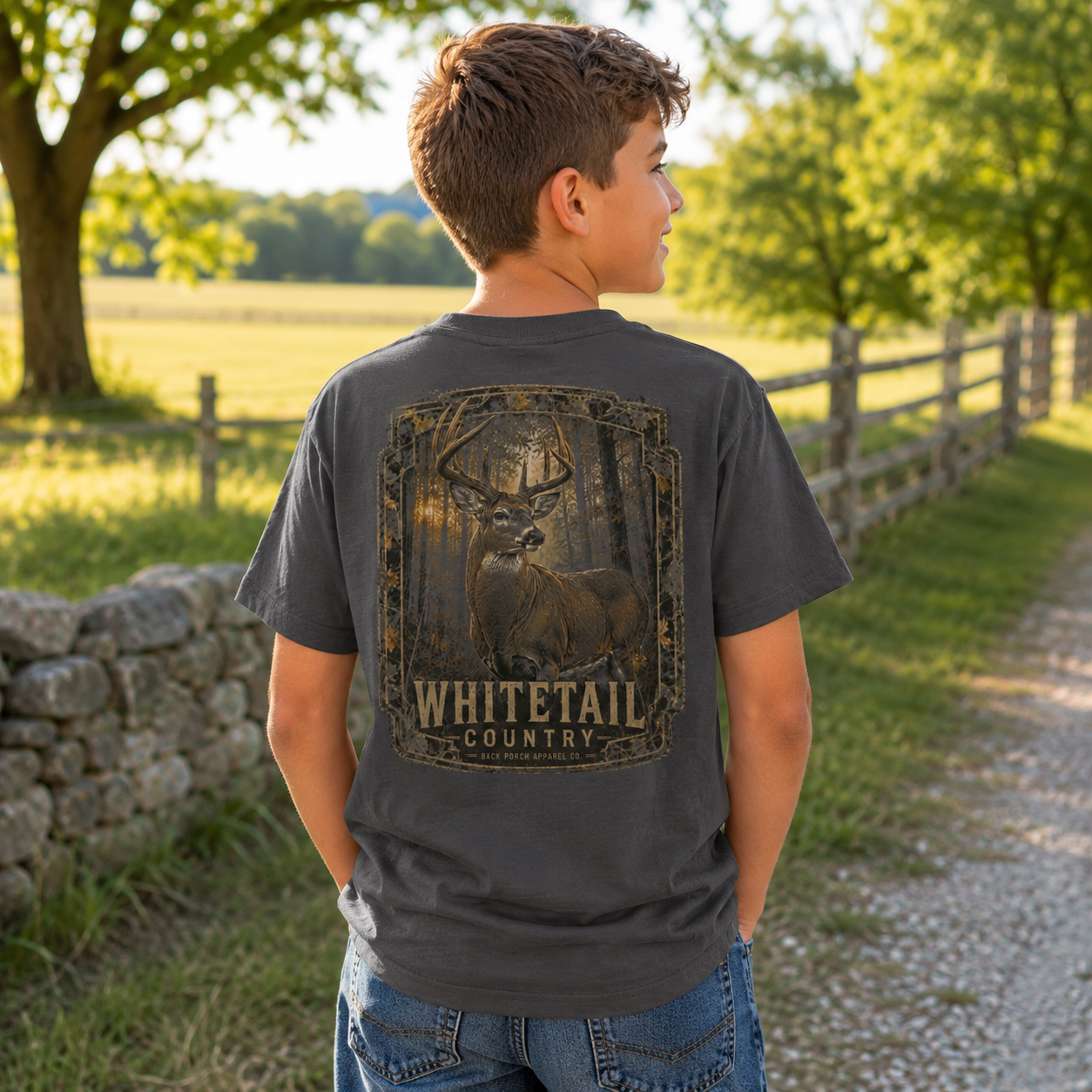 A boy standing outdoors on a country path wearing a dark gray t-shirt with a large whitetail deer graphic on the back and the words 'WHITETAIL COUNTRY' printed below it.