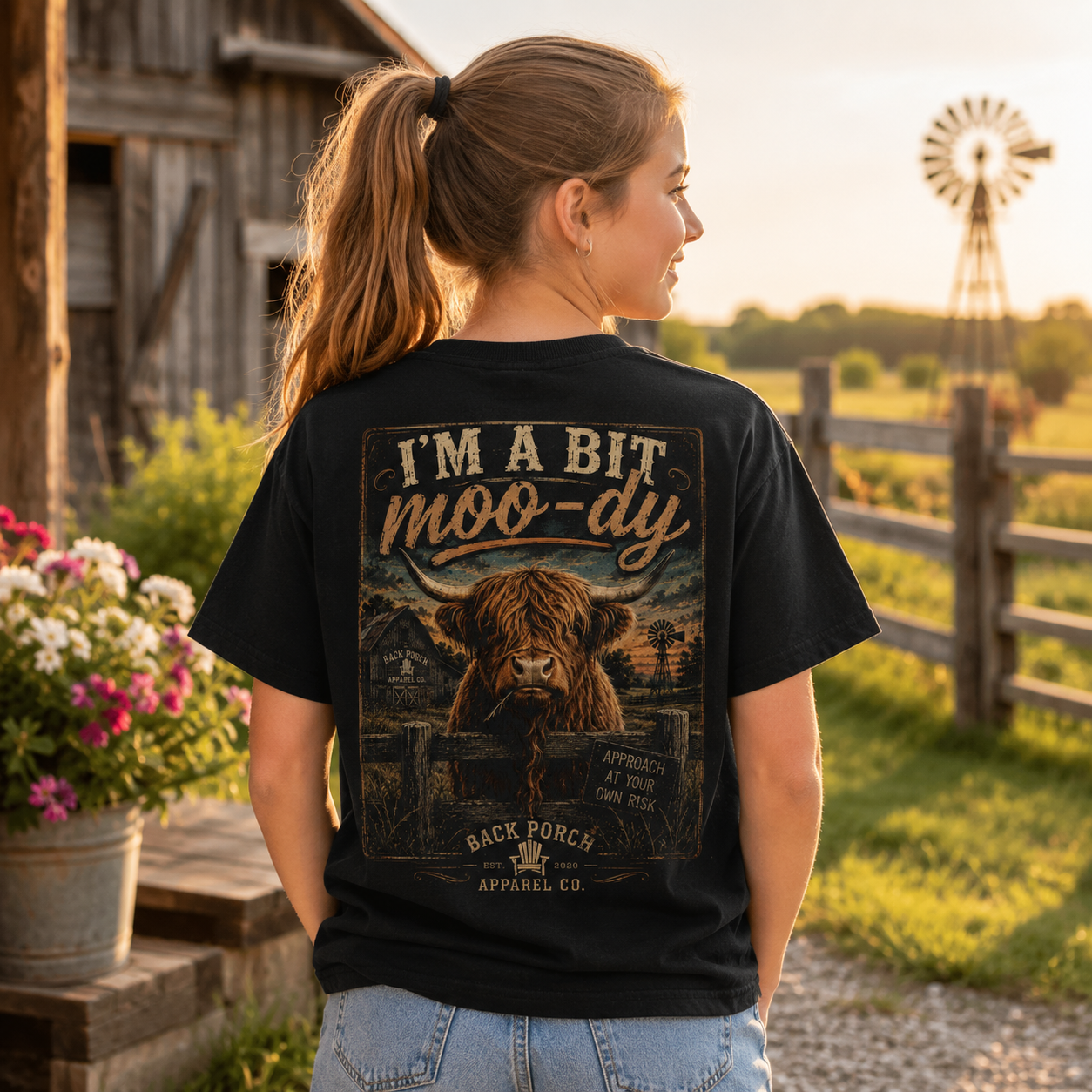 A woman stands outdoors on a farm wearing a black graphic T-shirt. The back of the shirt features a large vintage-style illustration of a highland cow and the text “I’M A BIT moo-dy” with “Back Forc Apparel Co.