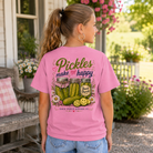 A woman wearing a pink graphic t-shirt with a pickle-themed design, standing on a porch in front of a house with greenery.