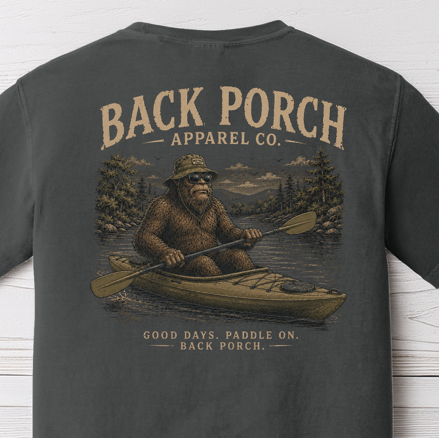 Bigfoot kayaking through a scenic mountain river illustration with Good Days Paddle On slogan by Back Porch Apparel