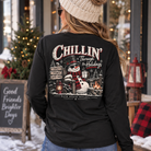 A woman viewed from behind wearing a black long-sleeve shirt with a large holiday snowman graphic on the back. The graphic reads “Chillin’ Through the Holidays” and shows a snowman with a red plaid scarf, lanterns, and a campfire.