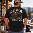 A man wearing a black graphic t-shirt with a large Santa Squatch design on the back, standing on a rustic outdoor porch with mountains and a fire pit in the background.