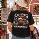 A woman viewed from behind wearing a black graphic t-shirt with a Highland cow and Christmas-themed text, set against a snowy outdoor holiday background.