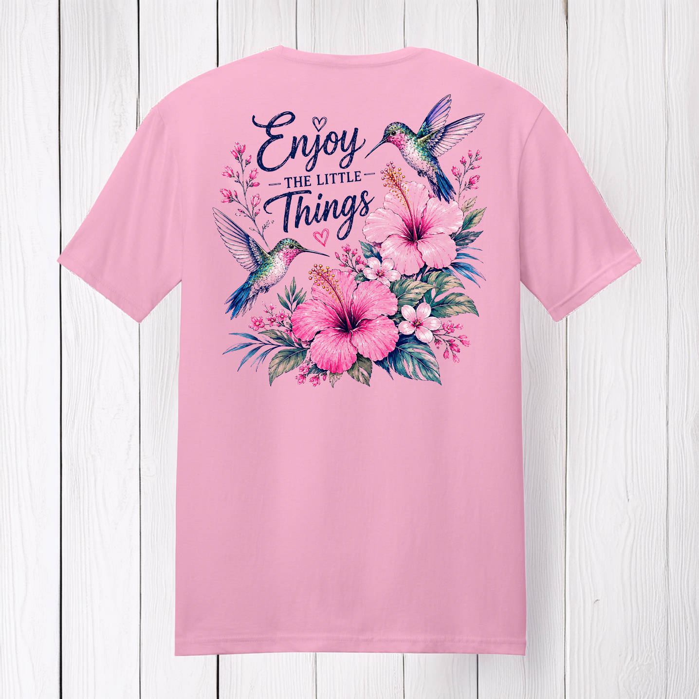 Enjoy The Little Things graphic t-shirt featuring colorful hummingbirds, pink flowers, and floral artwork on a soft pink shirt