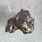 A rough, irregular piece of polished silver metal, resembling a small ingot or raw chunk, shown on a textured light gray background.