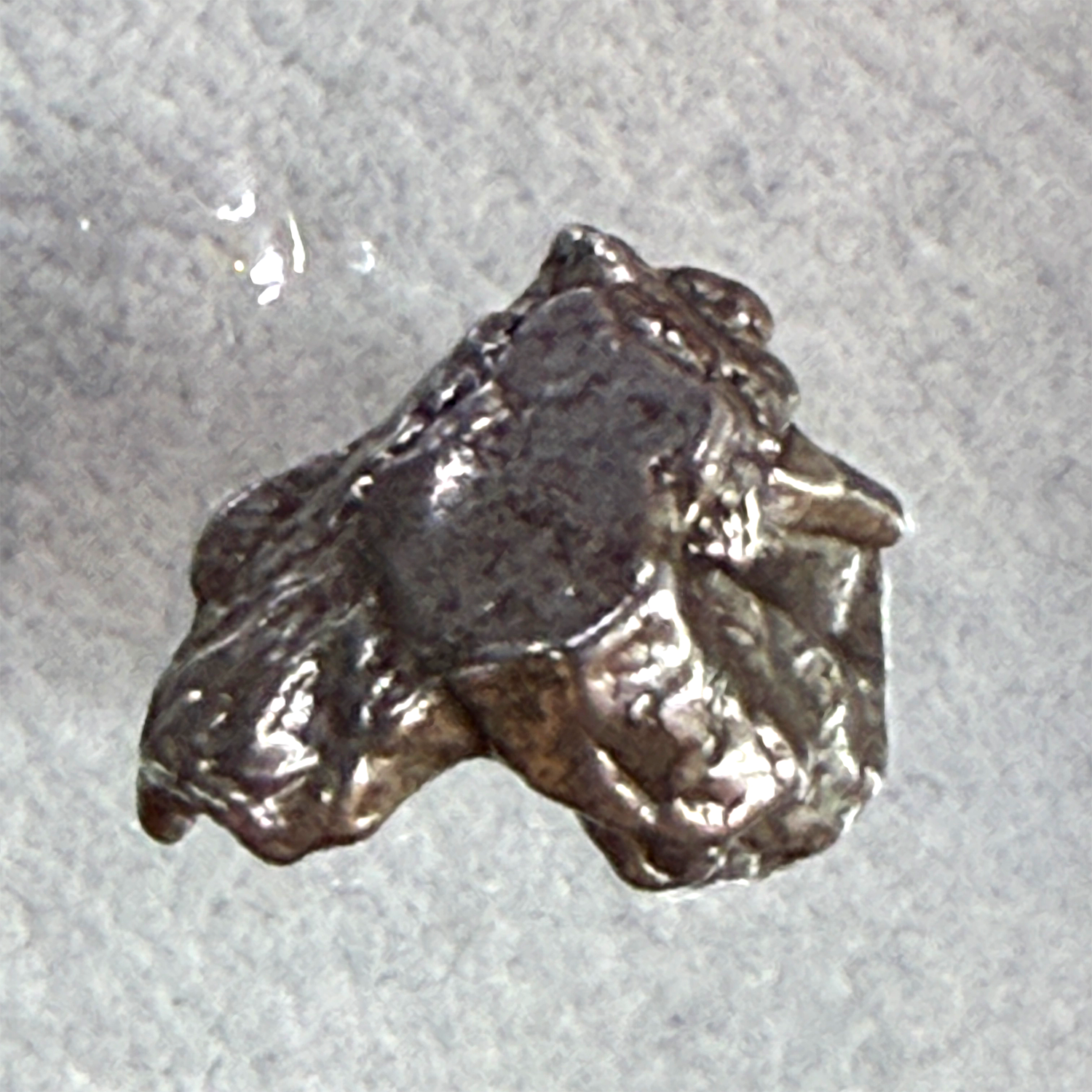 A rough, irregular piece of polished silver metal, resembling a small ingot or raw chunk, shown on a textured light gray background.