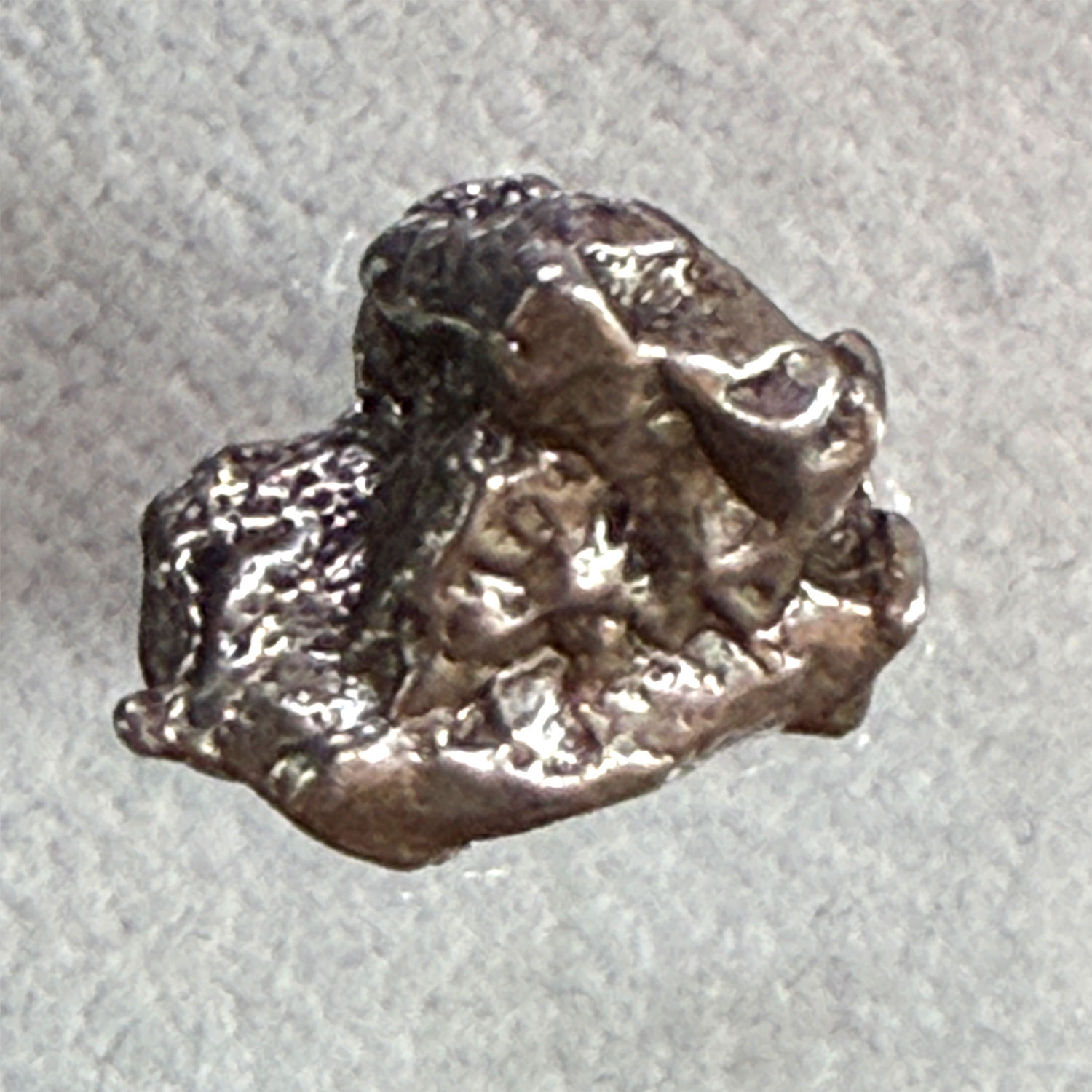 A small, rough, metallic lump of silver-colored metal, resembling raw sterling silver or pewter, photographed on a textured light gray background.