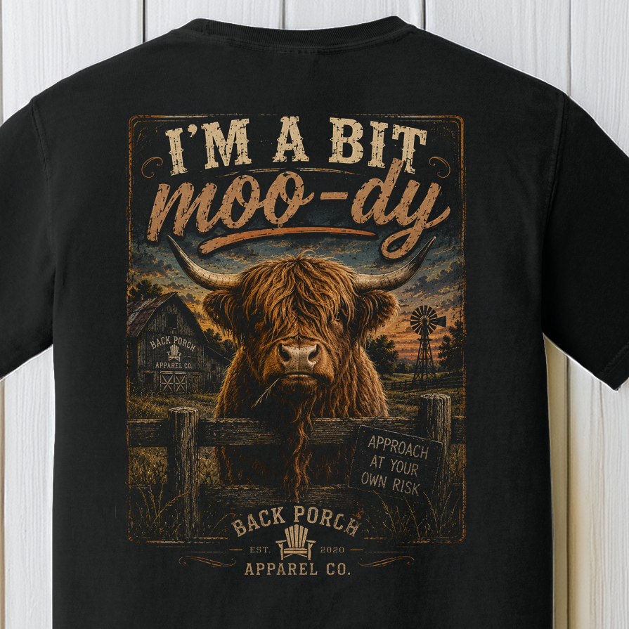 I'm A Bit Moo-dy Highland cow graphic t-shirt on Comfort Colors garment-dyed tee featuring a shaggy Highland cow behind a fence with country farm artwork