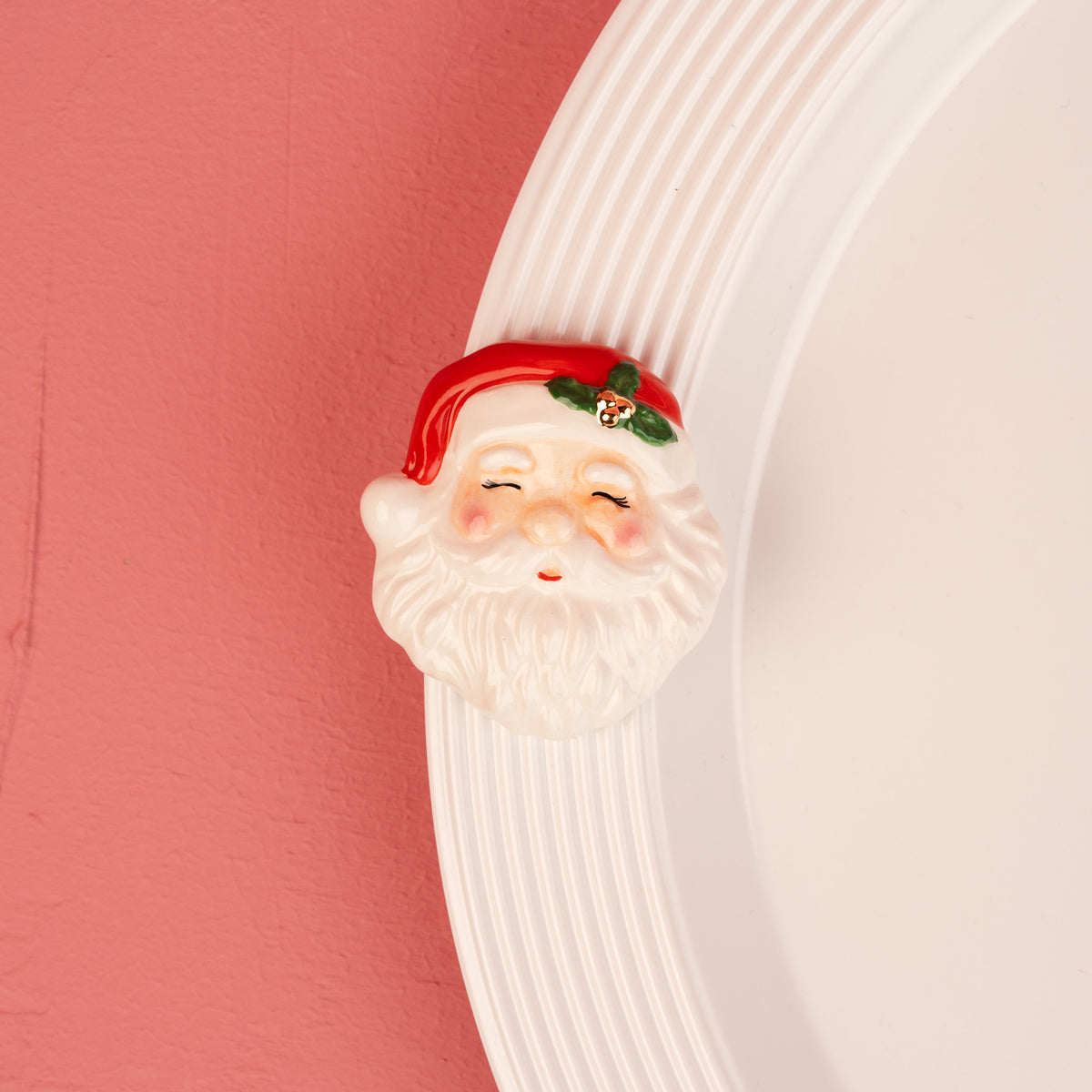A ceramic Santa Claus head wall hook with a red hat and white beard, mounted on a white ribbed circular plate against a pink wall.